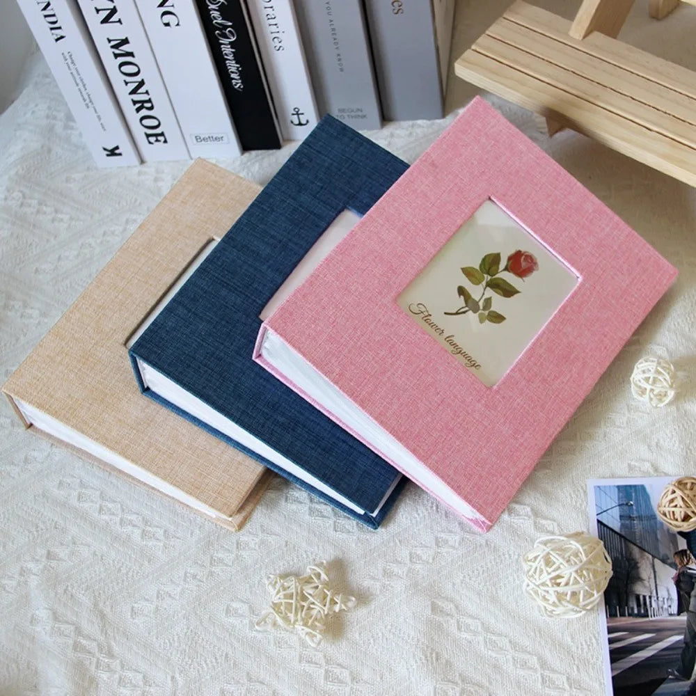A pink linen cover photo album with a floral design is displayed next to several books on a white textured surface.