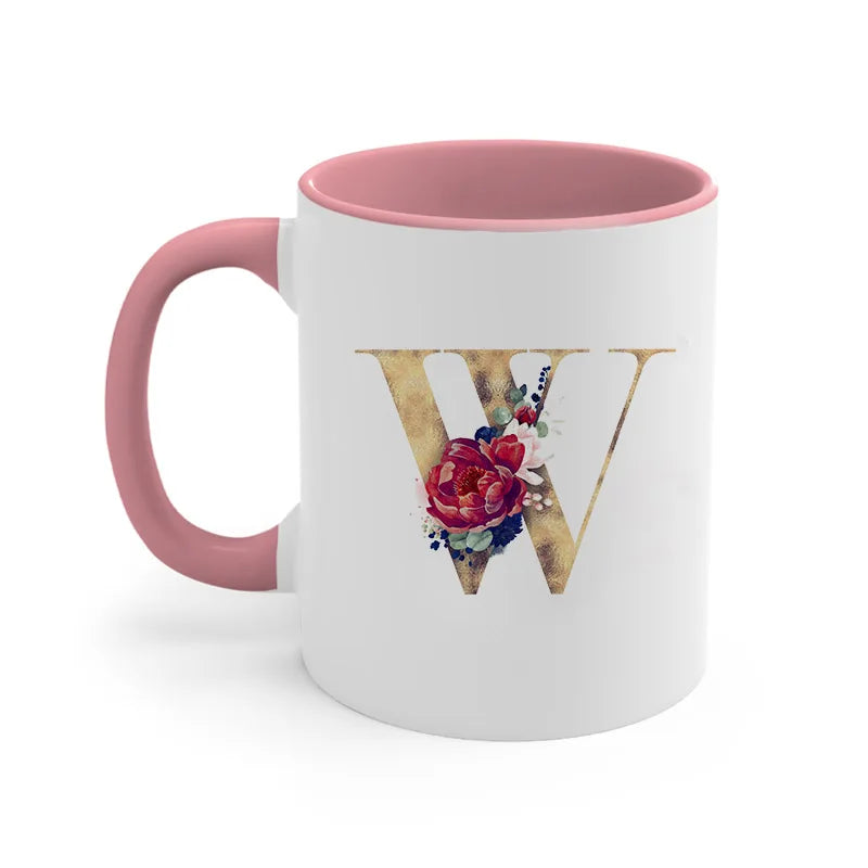A pink ceramic coffee mug featuring a gold letter W decorated with colorful flowers and greenery.