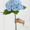 A cluster of light blue hydrangea silk flowers on a green stem and leaf, shown against a white background.
