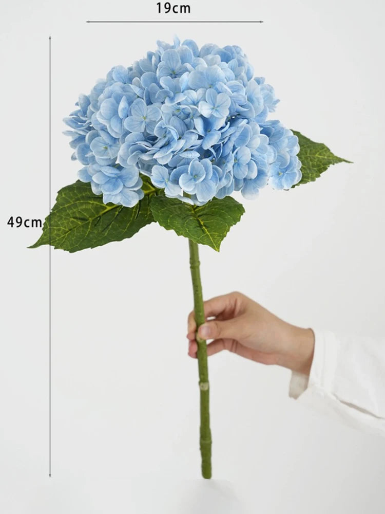 A cluster of light blue hydrangea silk flowers on a green stem and leaf, shown against a white background.