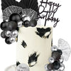 A white cake decorated with black and silver feathers and gold accents, topped with a black birthday banner and surrounded by