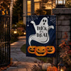 A dark blue flag featuring a white ghost silhouette and carved pumpkins displayed on a stone pillar in a garden setting.
