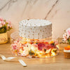 A white cake decorated with gold accents sits on a clear acrylic display stand filled with pink and yellow floral petals.