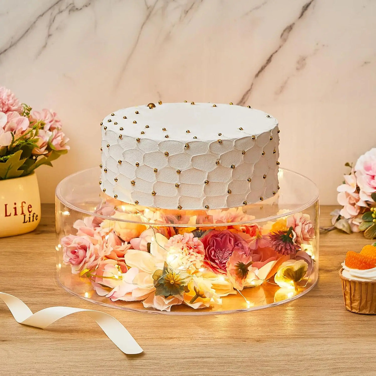 A white cake decorated with gold accents sits on a clear acrylic display stand filled with pink and yellow floral petals.