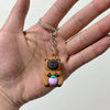 Un petit pendentif de capybara brun avec des accents violets est tenu dans la main d'une personne.