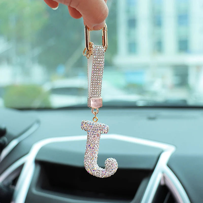 A hand holds a crystal letter keychain charm shaped like the letter J against a blurred background of a car interior.
