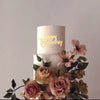 Weißer Kuchen mit goldenen Buchstaben 'Happy Birthday', dekoriert mit rosa und grünen Blumenakzenten.
