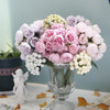 A bouquet of artificial pink peony silk flowers with pale purple and green accents in a glass vase.
