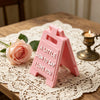 A pink 3D letter sign shaped like a house with the text A LITTLE SIGN TO SAY I LOVE YOU resting on lace fabric.