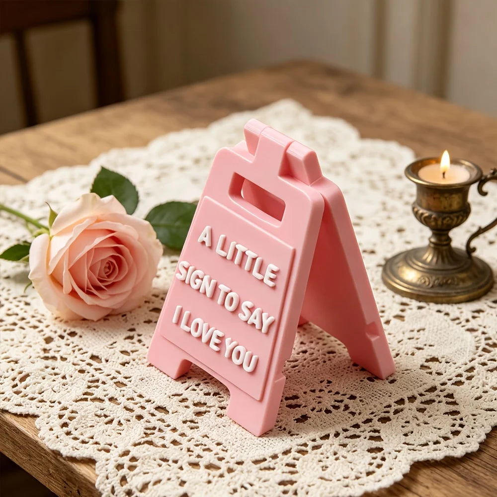 A pink 3D letter sign shaped like a house with the text A LITTLE SIGN TO SAY I LOVE YOU resting on lace fabric.