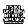 Black enamel pin with white text that reads It's not easy being my wife's arm candy but here I am nailing it