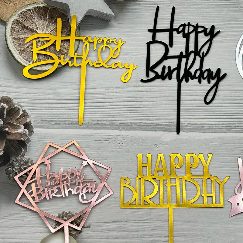 A set of acrylic birthday cake toppers in pink and gold lettering on a light gray wooden background.