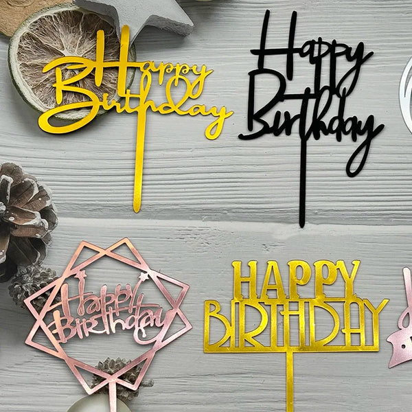 Un juego de toppers de pastel de cumpleaños de acrílico en letras rosa y dorado sobre un fondo de madera gris claro.