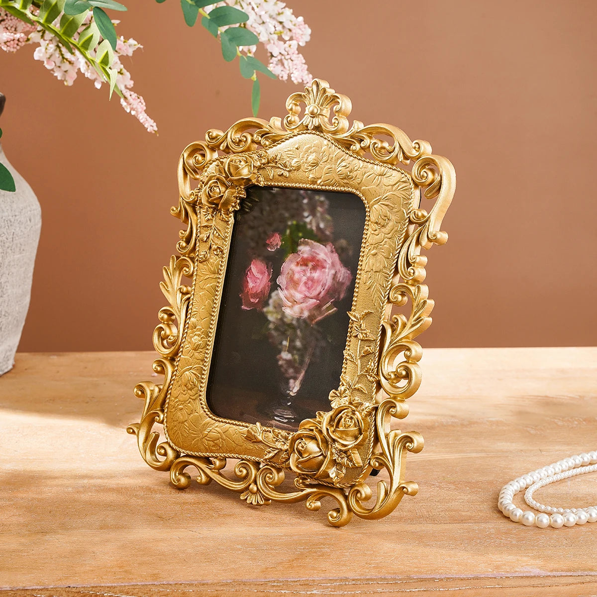 A gold-framed resin photo frame featuring a pink rose, displayed on a wooden surface.