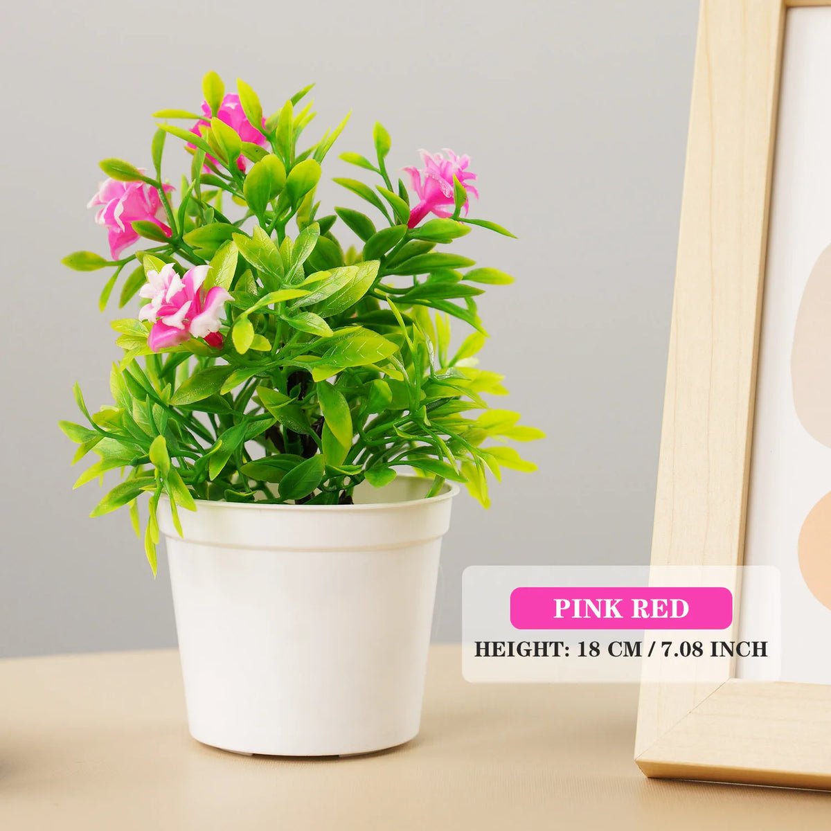 A potted artificial pink and green bonsai tree in a white pot displayed next to a wooden frame.