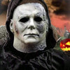 A close-up of a realistic Michael Myers horror costume mask with a pale, scarred face and dark hood.
