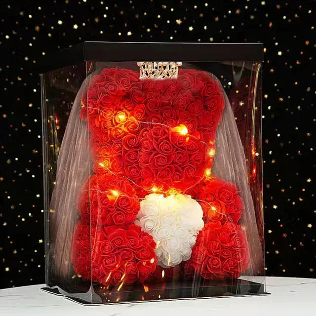 A red artificial rose teddy bear with white accents displayed in a clear box against a dark background.