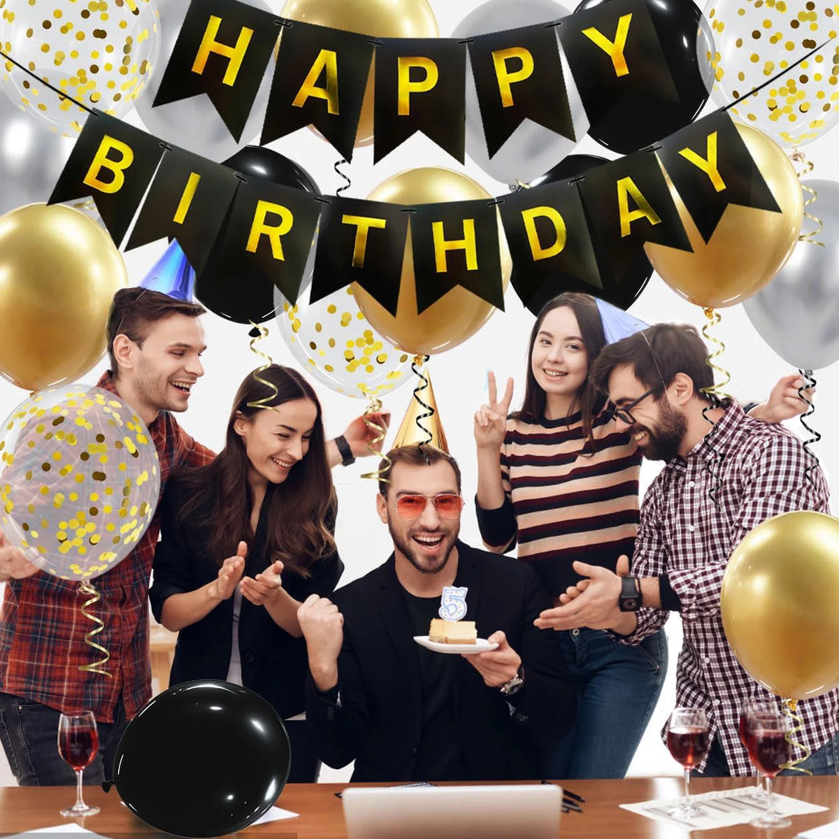 A group of people celebrating a birthday with black and gold balloons and banners.