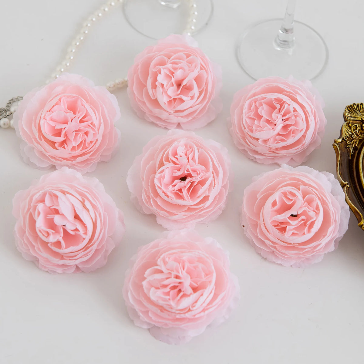 A collection of pink silk peony artificial flowers arranged on a white surface with a pearl necklace and gold object.