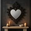 A dark metal ornate heart-shaped wall mirror surrounded by dark paneling and lit candles on a wooden shelf.