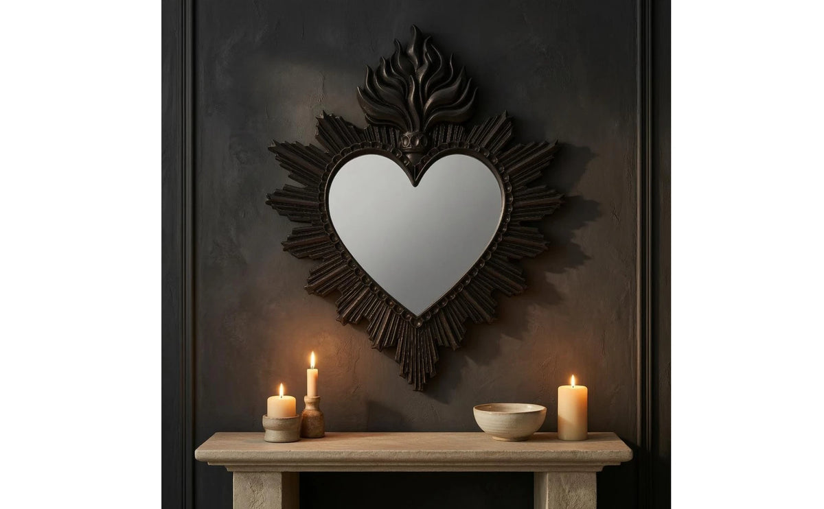 A dark metal ornate heart-shaped wall mirror surrounded by dark paneling and lit candles on a wooden shelf.