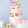 A tiered pink cake decorated with a castle topper, floral accents, and decorative elements for a birthday party.