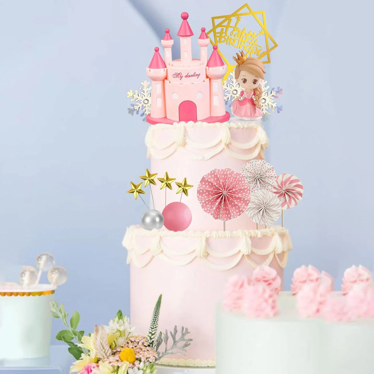 A tiered pink cake decorated with a castle topper, floral accents, and decorative elements for a birthday party.