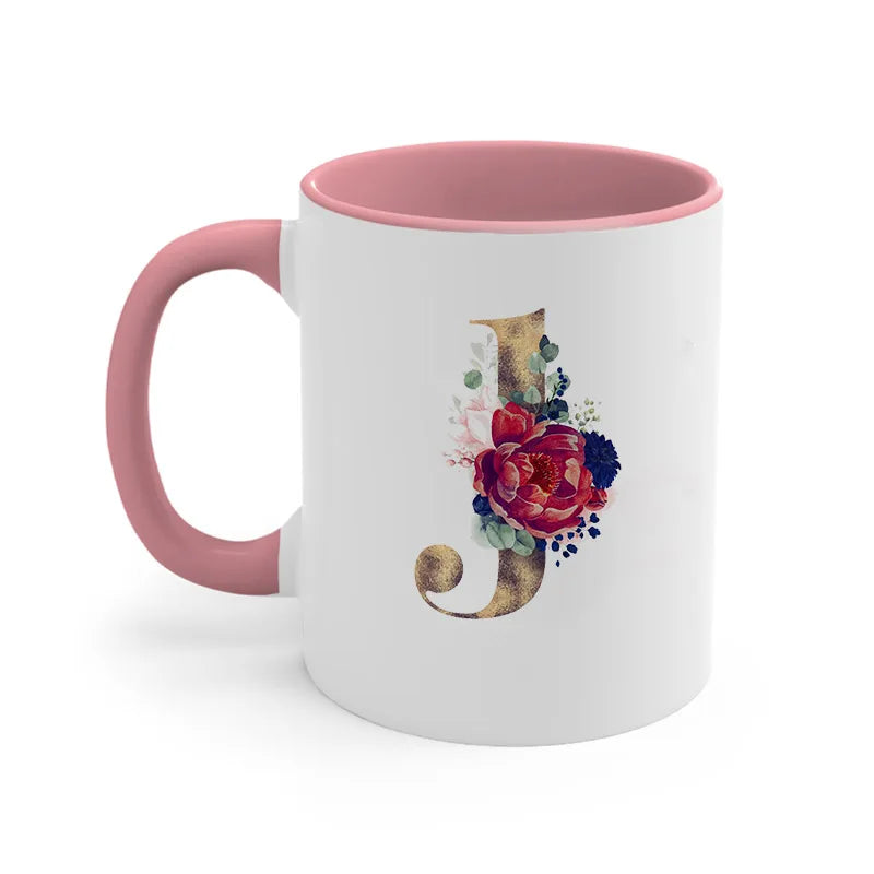 A pink ceramic coffee mug featuring a gold letter J design with floral accents.