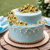 A two-tiered light blue cake decorated with gold ball toppers and small white flowers on a wooden surface.