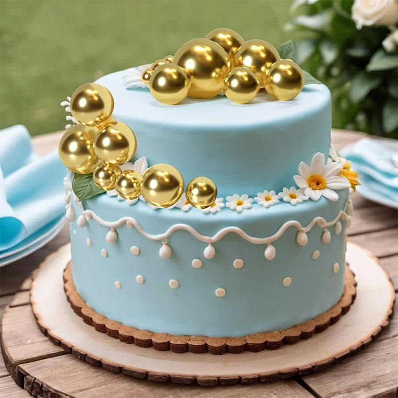 A two-tiered light blue cake decorated with gold ball toppers and small white flowers on a wooden surface.