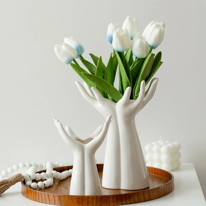 A white ceramic hand vase holds a bouquet of blue and white tulips on a wooden tray.