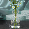 A glass vase holds a bouquet of blue and yellow flowers, set against a muted teal background.