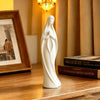 A white praying angel figurine statue standing on a wooden surface next to a framed picture and books.