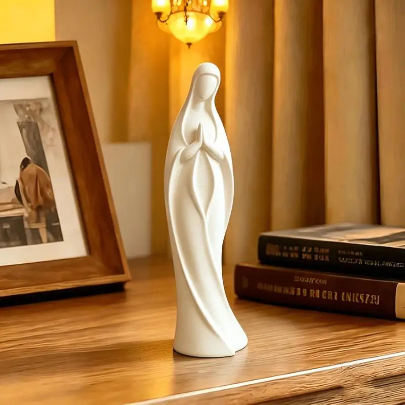 A white praying angel figurine statue standing on a wooden surface next to a framed picture and books.