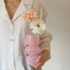 A person holds a pink glass flower vase with white floral arrangements against a white shirt.
