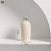 A beige minimalist ceramic vase with a round body is displayed on a white shelf.