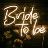 A neon sign spelling 'Bride to be' illuminates white roses against a dark, textured background.