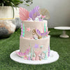 A tiered cake decorated with pink, purple, and green mermaid-themed toppers including fins, shells, and seaweed on a green su