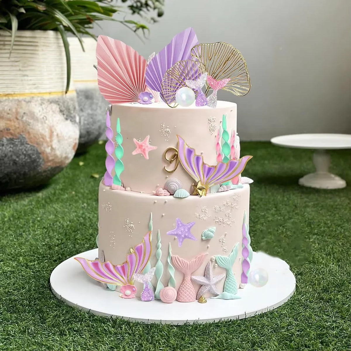 A tiered cake decorated with pink, purple, and green mermaid-themed toppers including fins, shells, and seaweed on a green su