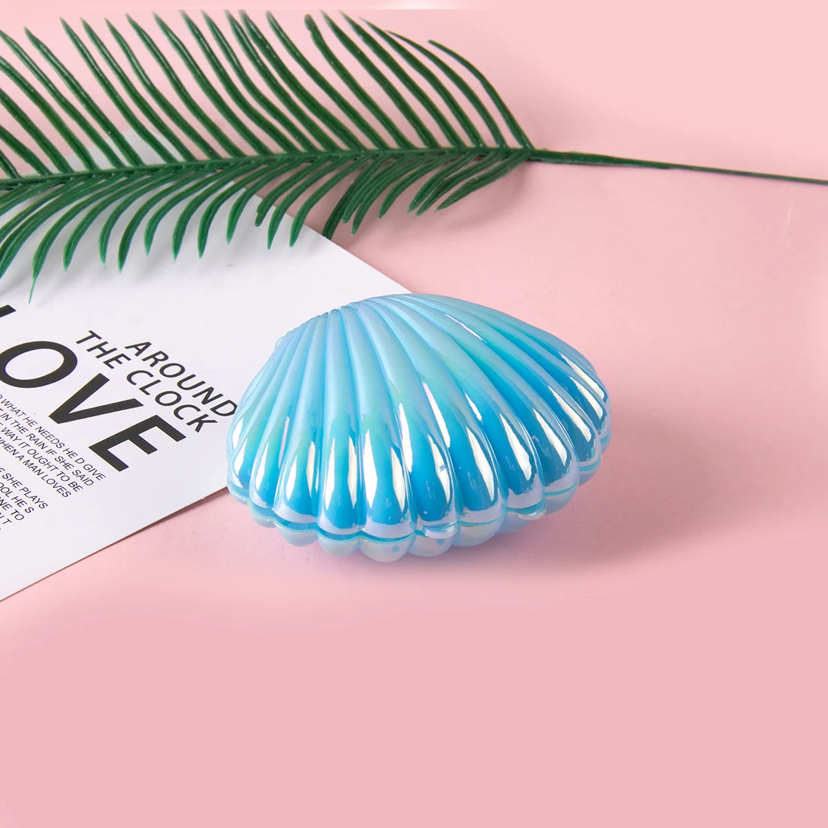 A translucent blue seashell resting on a pink surface next to a green palm frond.