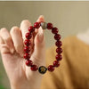 A hand is wearing a natural cinnabar stone zodiac bracelet with gold-colored beads and a dark central stone.