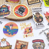 A collection of colorful graffiti stickers featuring characters and text like This Is An Outrage, Dungeon Crawler, and God Da