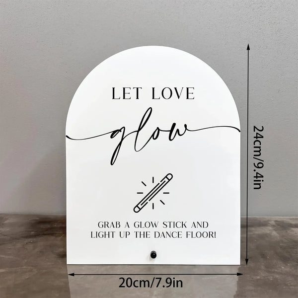 A white acrylic sign with black script text that reads Let Love glow and features an illustration of a glow stick.
