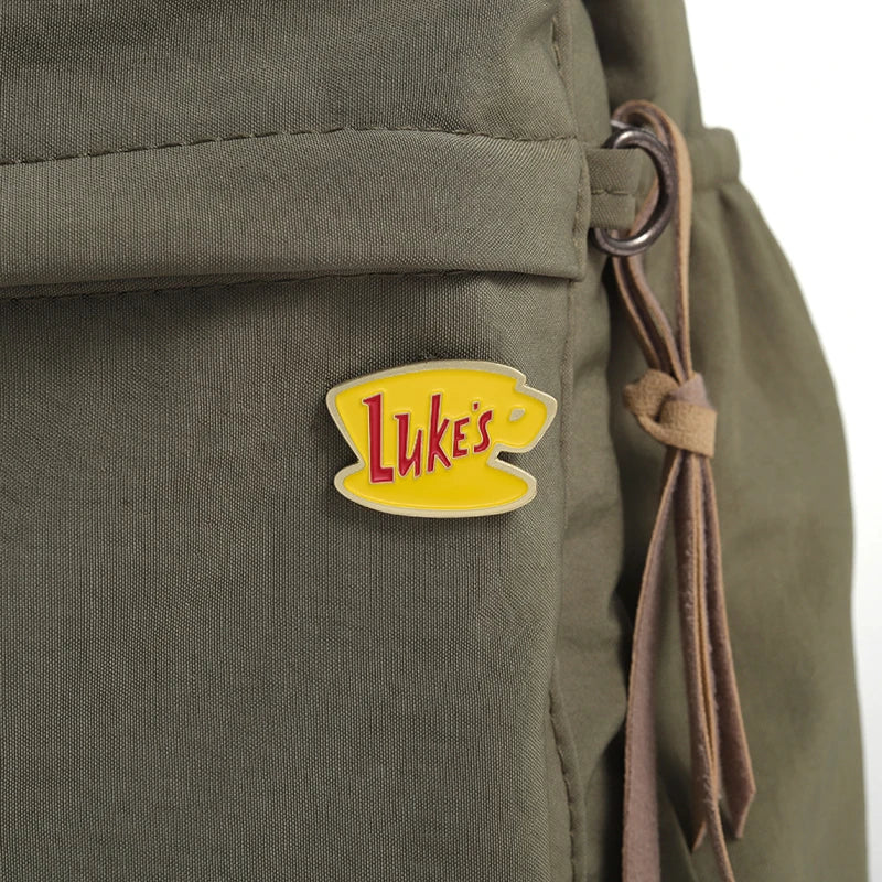 A yellow enamel pin shaped like a smiling face with the word Luke's on it attached to olive green fabric.