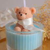 A fluffy orange teddy bear candle in a glass jar with a white ribbon, set against a blurred background.