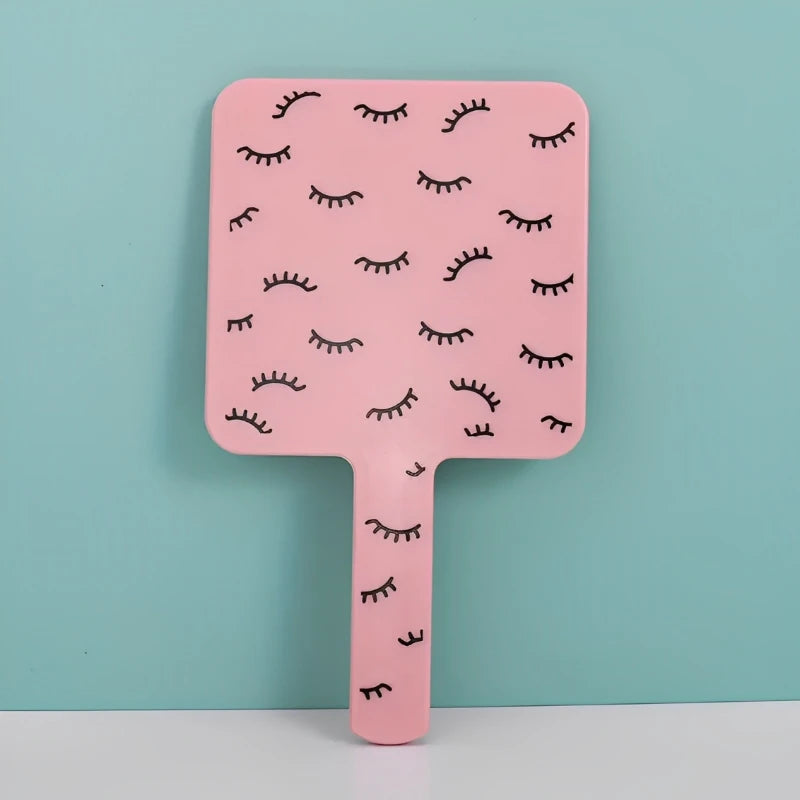 A pink handheld vanity mirror with an ergonomic handle featuring a pattern of stylized eyelashes.