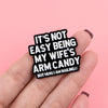 A hand holds a black enamel pin with the text It's not easy being my wife's arm candy but here I am nailing it