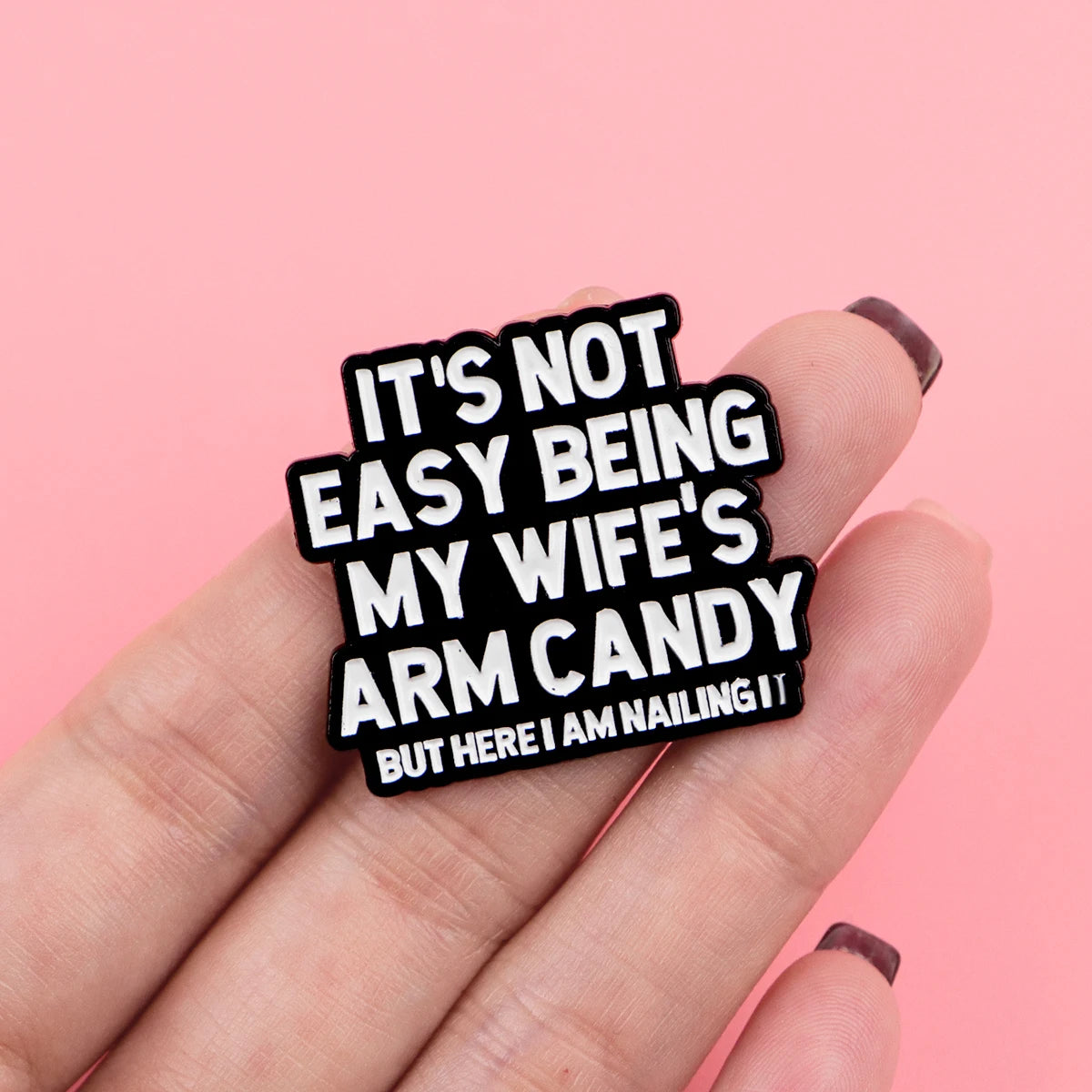 A hand holds a black enamel pin with the text It's not easy being my wife's arm candy but here I am nailing it