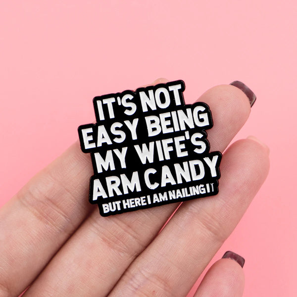 A hand holds a black enamel pin with the text It's not easy being my wife's arm candy but here I am nailing it
