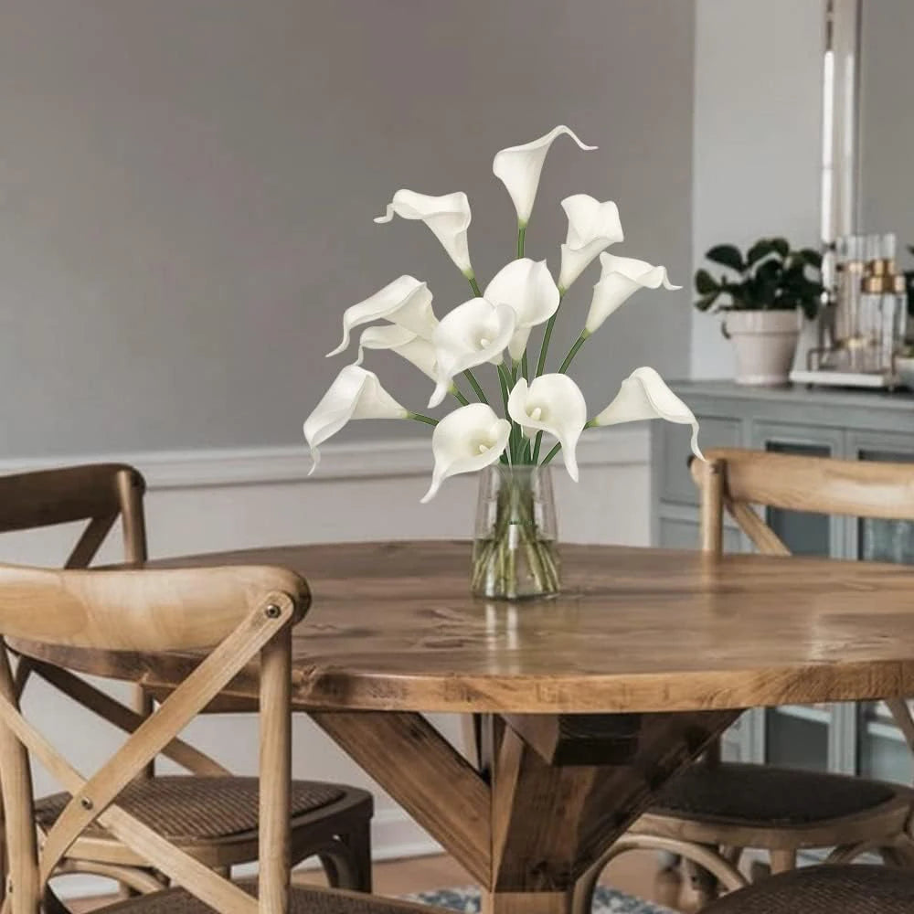 A glass vase holds a bouquet of white calla lily artificial flowers on a wooden table.
